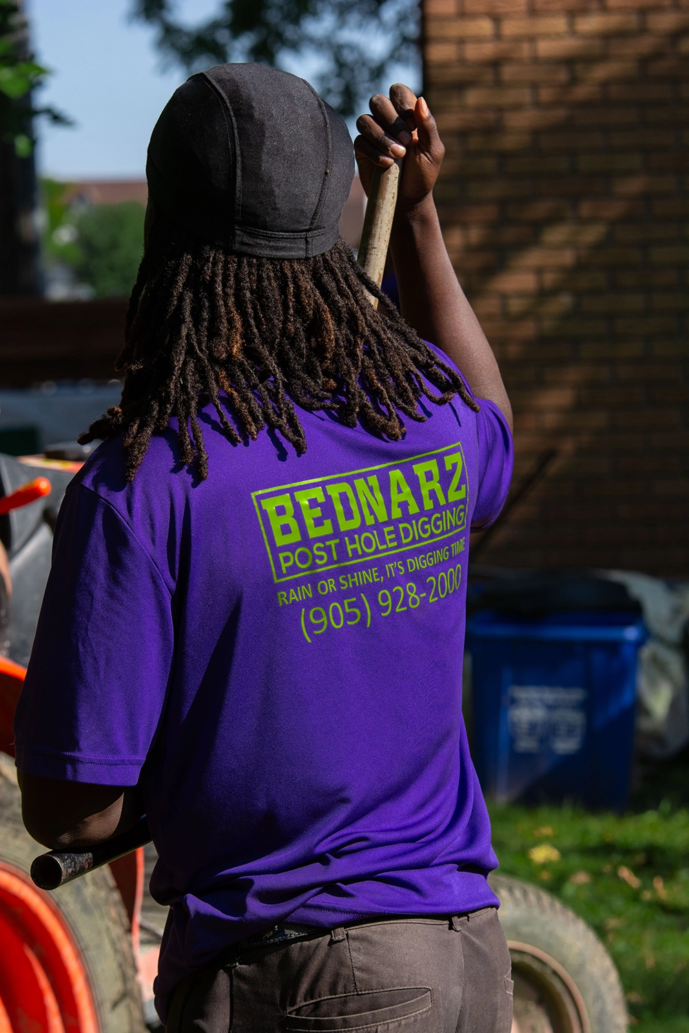 Bednarz crew member in purple branded shirt and cap carrying wooden post for fence installation at Paris Ontario residential site